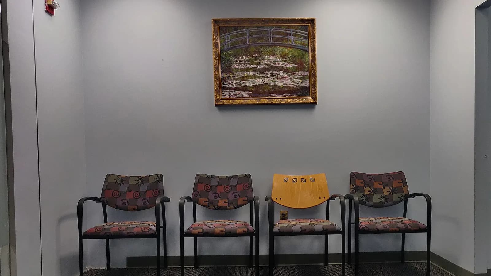 Sala d'attesa vuota di uno studio medico: quattro sedie imbottite contro una parete azzurra sotto un quadro con un ponte su uno stagno di ninfee (foto: Kurt Kaiser, CC0, Wikimedia Commons)